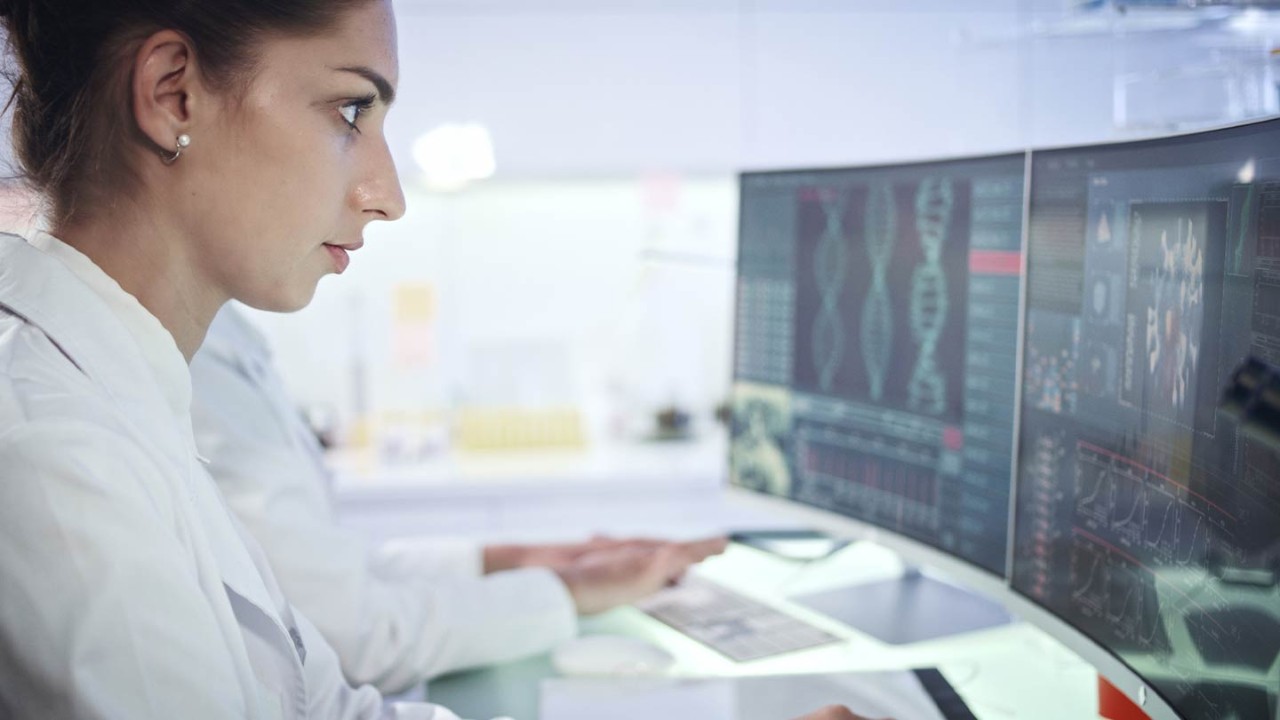 Woman healthcare professional sitting at desk, examining genetic data on a desktop monitor.