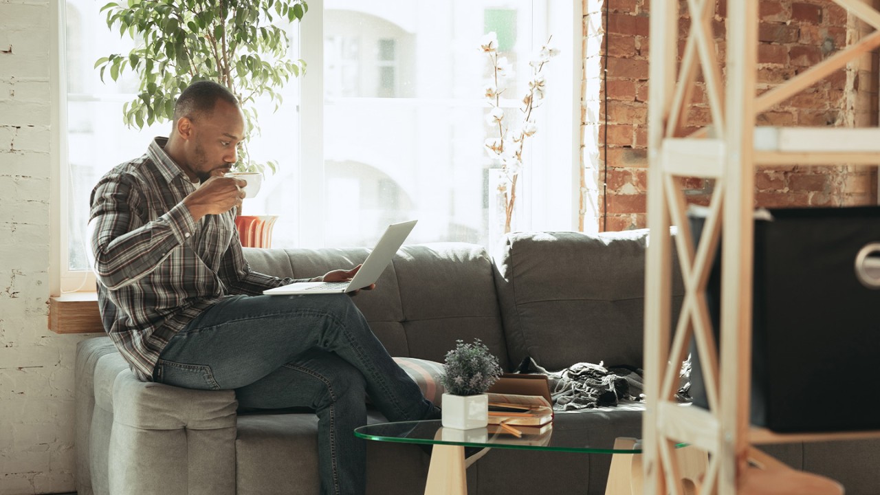 Man sitting on the arm of a couch with a coffee cup in his hand and a laptop on his lap.