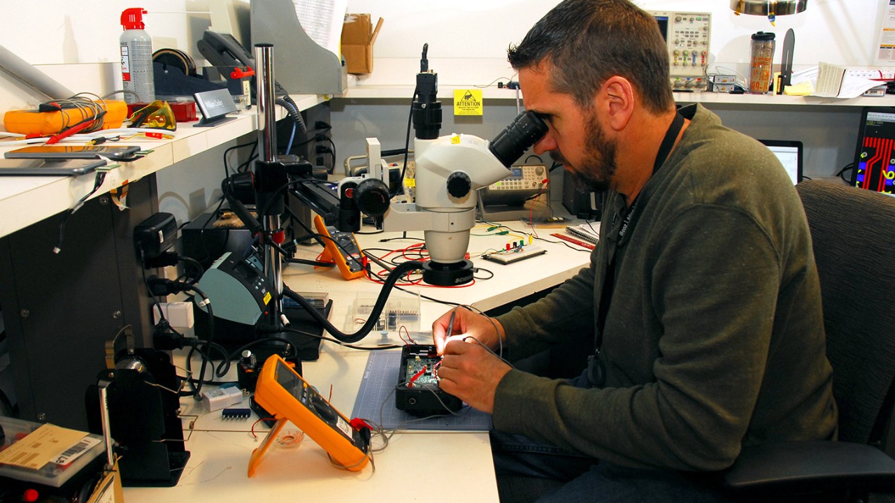 Engineer sitting at a desk looking through a microscopic lens building custom electronics.