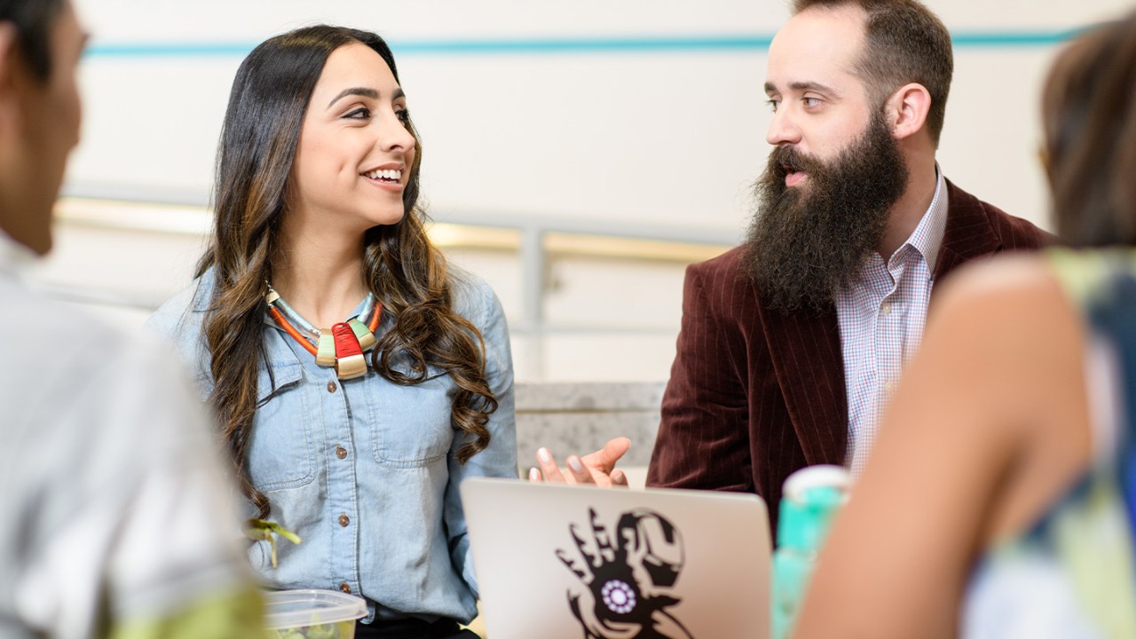 Woman sitting and talking with a bearded man looking a laptop on a table with other co-workers surrounding the table.