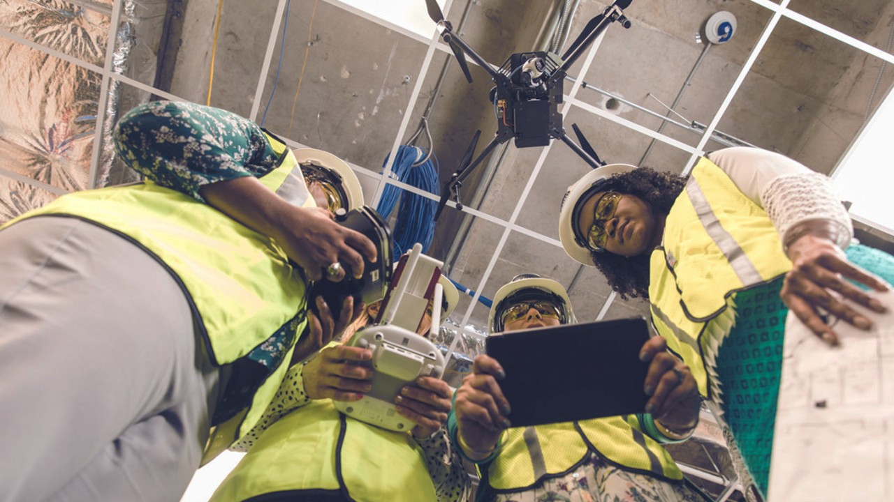 Four construction workers inside a building, outfitted in yellow vests and hard hats, with each person holding a piece of digital equipment.