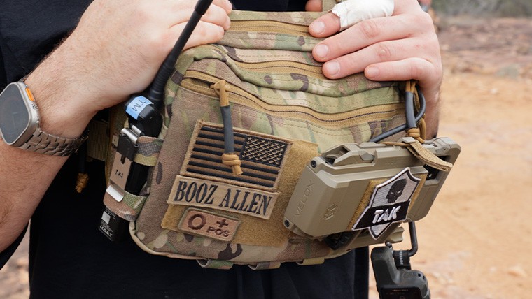 Close-up of a person wearing a multicam tactical chest pack featuring a Booz Allen patch, a radio, and a mounted Velox EUD device with a "TAK" skull patch.
