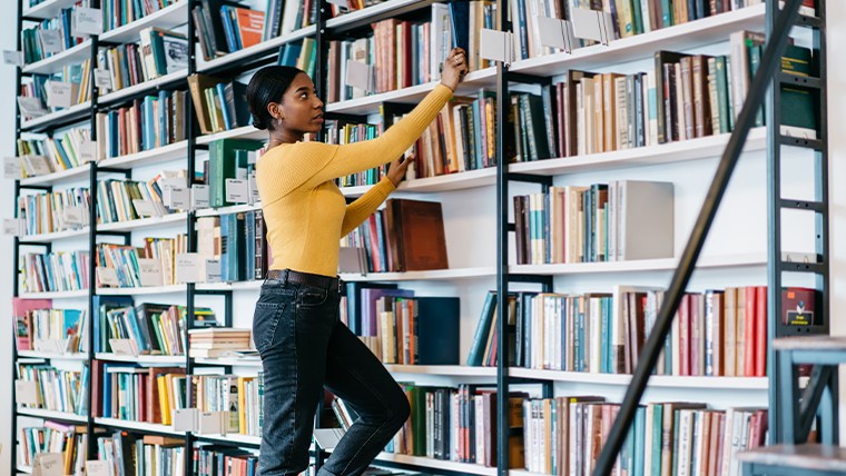 Woman on a ladder reaching for a book out of a bookcase. 