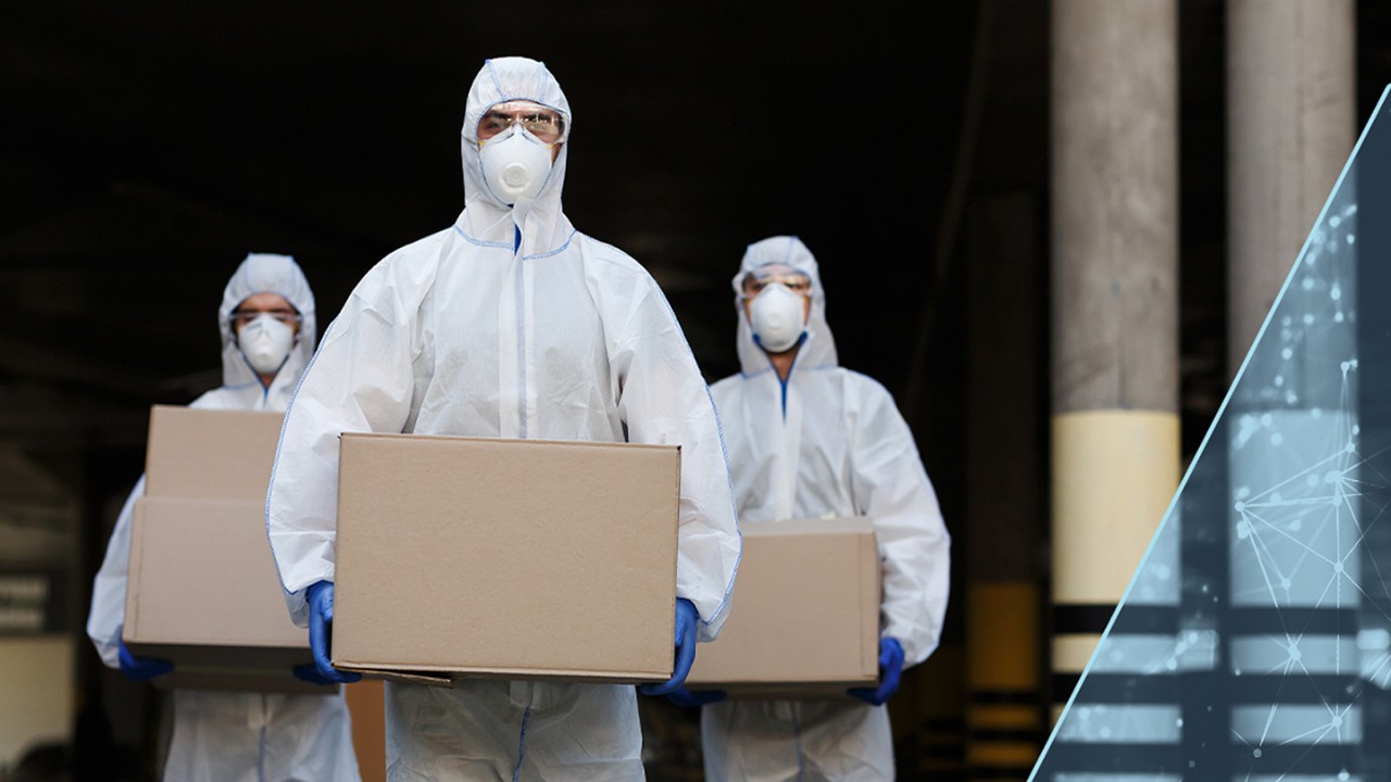 Three people in protective suits, goggles and masks, holding boxes of the vaccine exiting a warehouse.