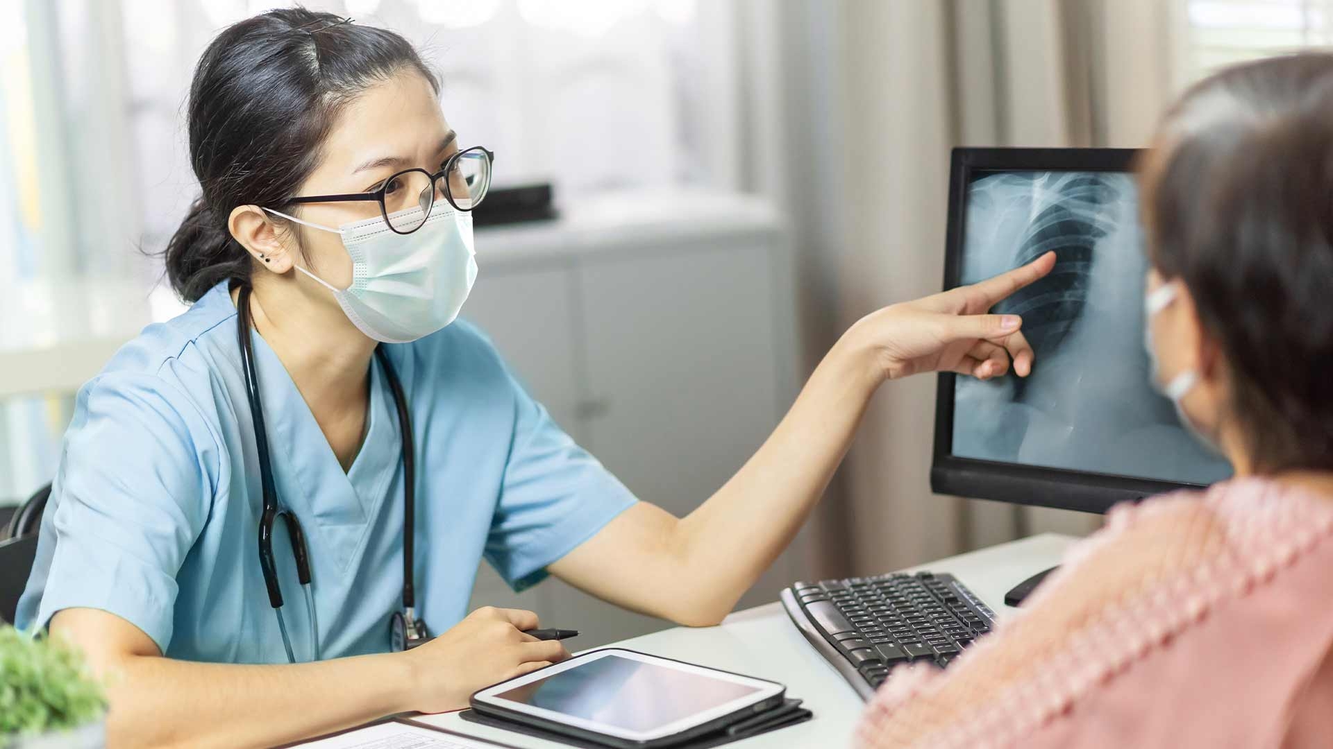 Female doctor in medical mask examining and pointing to radiological chest X-ray film on monitor computer while talking with elderly woman patient at hospital room.
