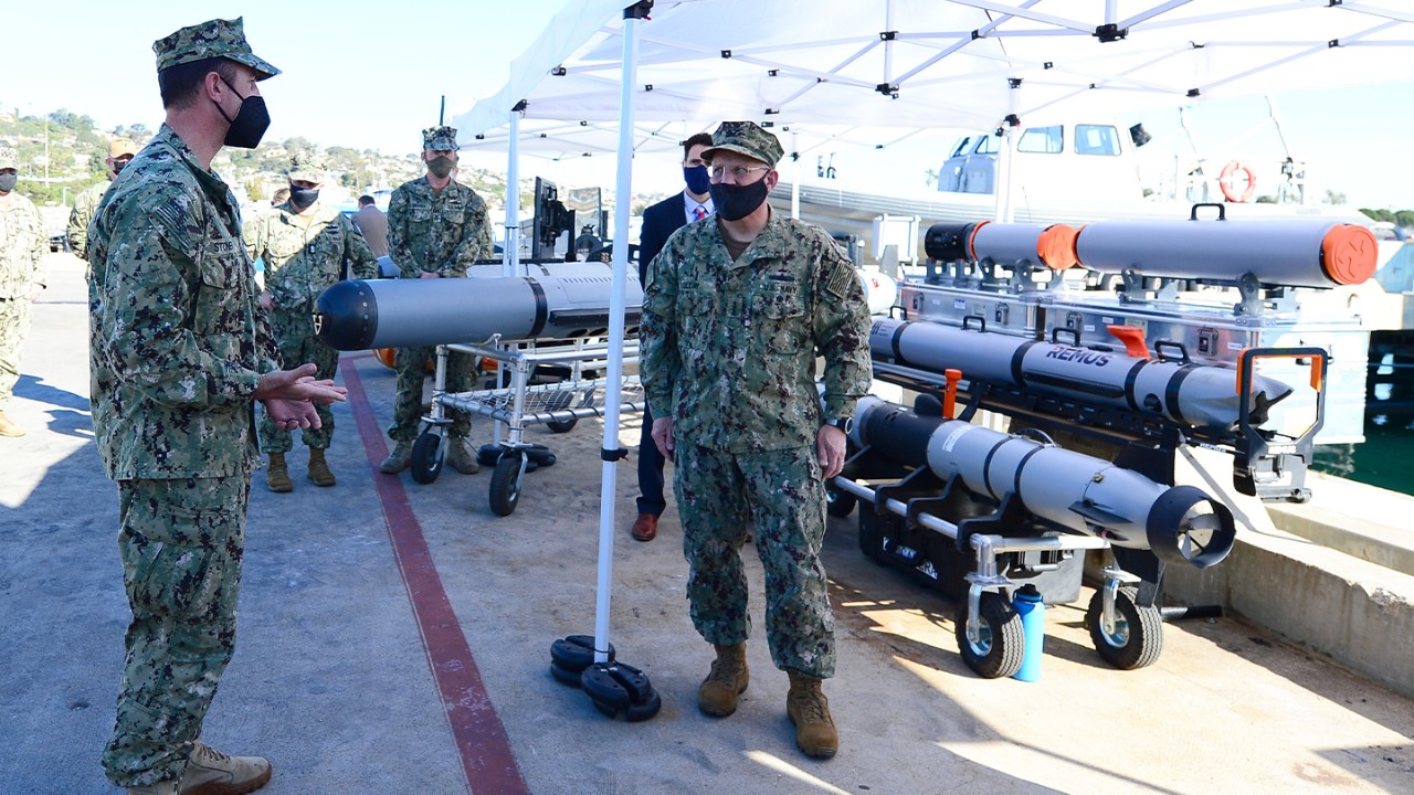 A group of warfighters talks in front of military weapons